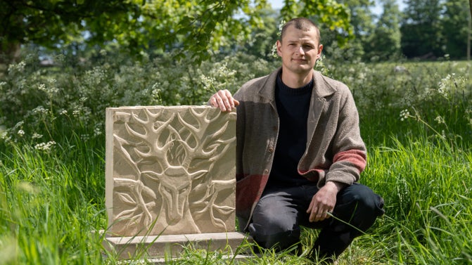 A man is crouched down in an area with long grass with his arm rested on a stone plinth. A deer's face is carved into the front of the stone.